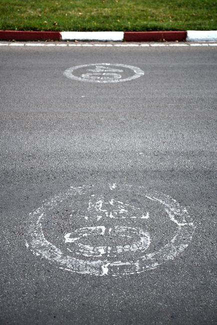 A photograph of a paved street showing two circular road markings painted in white. The markings are worn and partially faded; the one near the top of the image is smaller and cleaner, while the larger one closer to the bottom is more deteriorated with chipped edges. The street surface is smooth with visible small cracks and debris. A curb with red and white painted stripes runs along the top edge, separating the road from a grassy area. The grassy strip appears well-maintained with short, green grass. There are no vehicles, people, or additional objects visible. This scene could be related to street regulation or parking considerations for house removals or moving services, such as those provided by Man with Van Tottenham Hale, involved in furniture transport and home relocation, especially when adhering to local pavement loading rules during a handover or loading process.