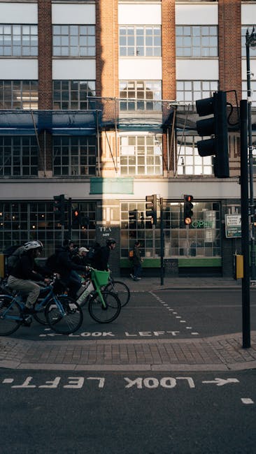 The image shows a street scene in front of a multi-storey brick building with large windows. Several people are riding bikes along the road, with some wearing helmets. A group of pedestrians is walking on the sidewalk, passing a commercial unit with glass doors and windows. A traffic light is visible at the corner, mounted on a black pole with pedestrian crossing signals. The pavement includes a designated loading and unloading zone marked with white text and arrows, indicating 'LOOK LEFT.' The scene suggests urban activity during daylight hours, with natural light illuminating the area. This setting aligns with the context of house removals and moving logistics, relevant to the topic of pavement loading rules for movers in Tottenham Hale as discussed on manwithvantottenhamhale.co.uk.