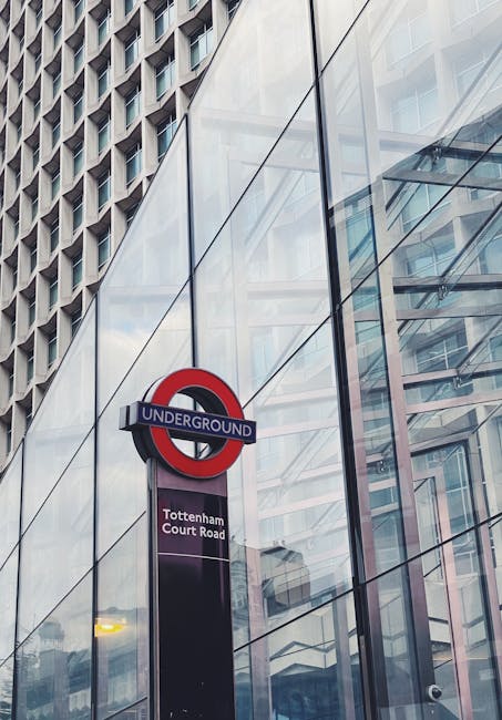 A tall, modern glass building with reflective panels showcasing the surrounding urban environment is visible in this image. Mounted on a black pole in front of the building is a round London Underground sign with a red circle, blue bar, and white lettering, indicating the Tottenham Court Road station. Below the sign, there is a rectangular sign displaying the station name. The scene is captured during daylight hours with natural light illuminating the area, highlighting the clean, sleek lines of the glass surfaces. The reflection on the glass panels shows a partial view of nearby structures, contributing to the cityscape atmosphere. This urban setting relates to the context of house removals and relocation services, with Man with Van Tottenham Hale providing professional assistance in shifting belongings through busy city environments.