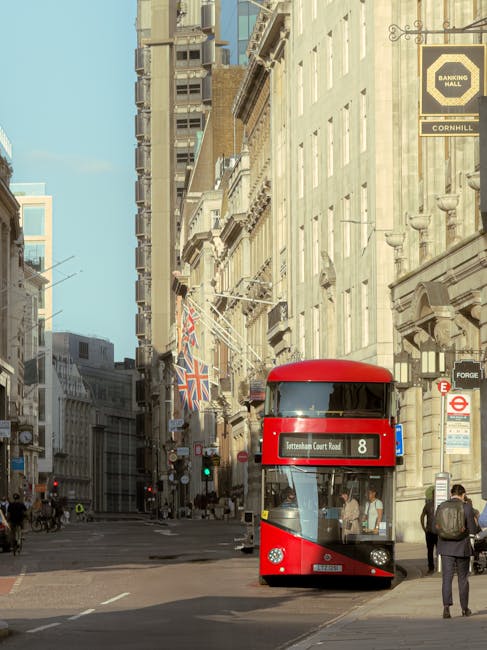 A tall, modern glass building with reflective panels showcasing the surrounding urban environment is visible in this image. Mounted on a black pole in front of the building is a round London Underground sign with a red circle, blue bar, and white lettering, indicating the Tottenham Court Road station. Below the sign, there is a rectangular sign displaying the station name. The scene is captured during daylight hours with natural light illuminating the area, highlighting the clean, sleek lines of the glass surfaces. The reflection on the glass panels shows a partial view of nearby structures, contributing to the cityscape atmosphere. This urban setting relates to the context of house removals and relocation services, with Man with Van Tottenham Hale providing professional assistance in shifting belongings through busy city environments.
