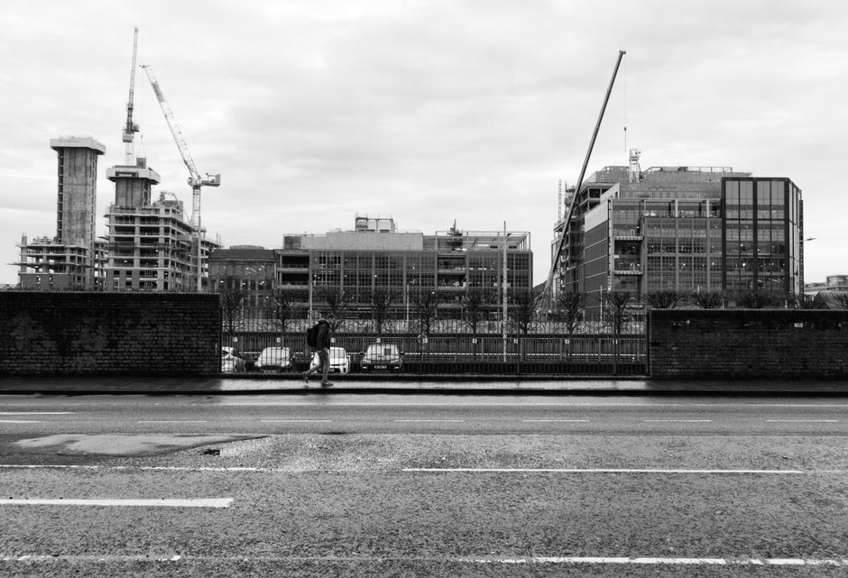 A black-and-white photograph showing the exterior of a modern, multi-storey building under construction in an urban area, with several cranes visible on the construction site. In the foreground, a str