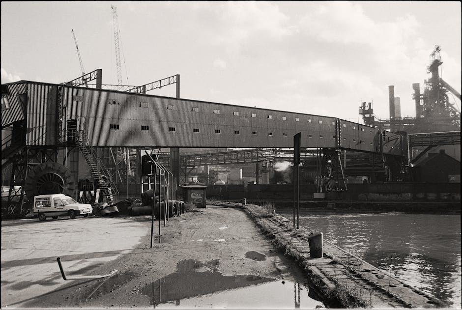 A black-and-white photograph showing the exterior of a modern, multi-storey building under construction in an urban area, with several cranes visible on the construction site. In the foreground, a str