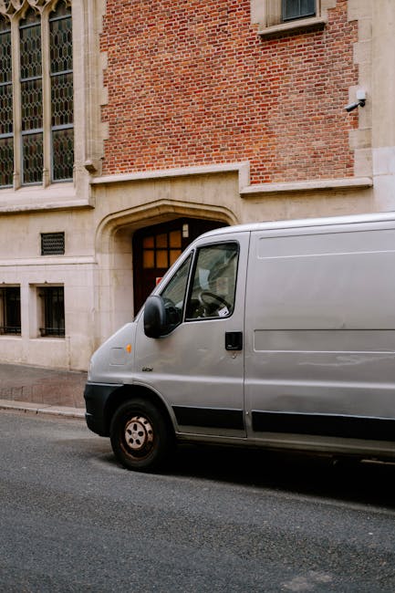 A silver removal van parked on a street in front of a red-brick building with large stained-glass windows and stone detailing around the entrance. The van's side door is closed, and it is positioned close to the pavement, with the front wheel turned slightly towards the curb. The surrounding environment appears quiet and urban, with no other vehicles or pedestrians visible. The scene depicts a typical home relocation or furniture transport scenario in progress, with the van ready to assist with moving household items, as part of a professional removals service. Man with Van Tottenham Hale's vehicle is stationary in a suitable location for loading or unloading furniture, boxes, and packed belongings, fitting with the context of street-level home moves or moving logistics near residential or institutional buildings.