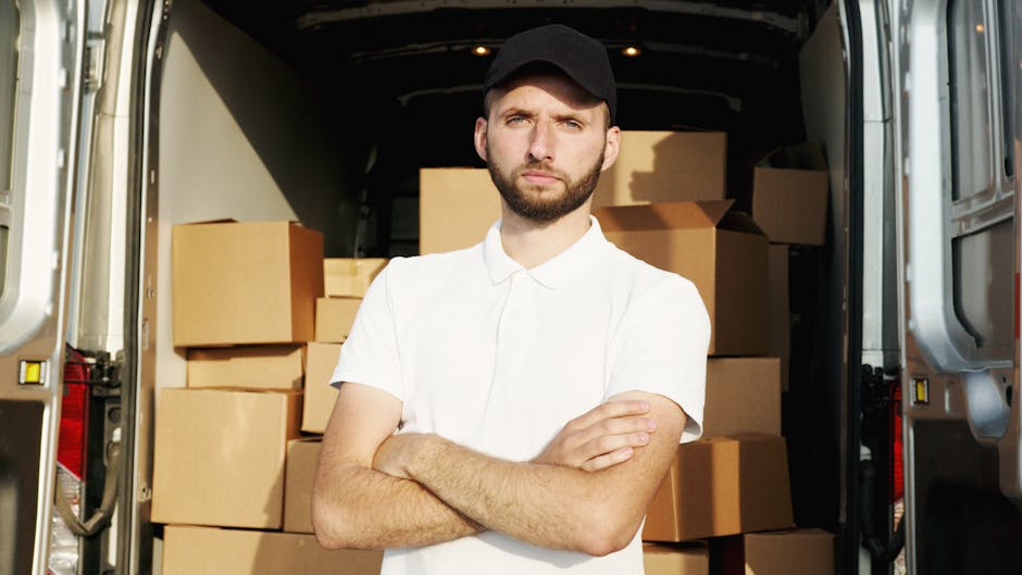 A man with a beard, wearing a black cap and a white polo shirt, stands inside the open rear of a van loaded with neatly stacked cardboard boxes. The boxes are various sizes, some wrapped in plastic, a