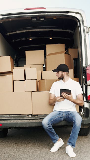 A man with a beard, wearing a black cap and a white polo shirt, stands inside the open rear of a van loaded with neatly stacked cardboard boxes. The boxes are various sizes, some wrapped in plastic, a