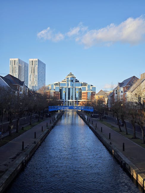 A photograph showing a canal lined with paved walkways and low black bollards on either side, with leafless trees along the edges. On the left, there are residential houses with brick walls and pitched roofs, while on the right, similar houses are visible. The canal leads to a modern office building with a distinctive, layered blue glass facade and a rounded top, situated in the background against a bright blue sky with a few white clouds. In the foreground, a professional removals team from Man with Van Tottenham Hale is loading furniture and packed boxes into a blue van parked on the right side of the pavement, with some items covered in protective plastic wrap and cardboard. The team is using trolleys, straps, and blankets to facilitate the home relocation and furniture transport process, reflecting typical packing and loading procedures during a domestic move in an urban setting.