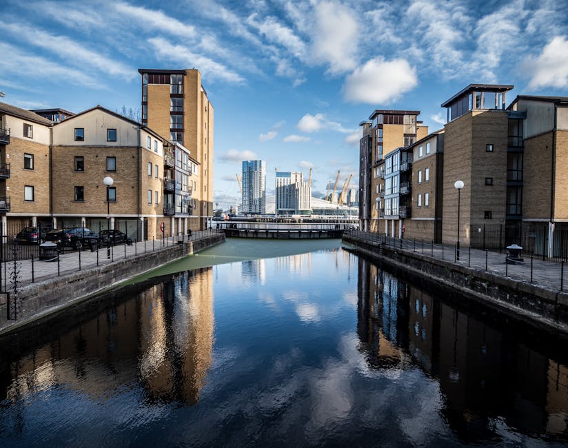 A daytime view of residential buildings on both sides of a narrow canal with calm water reflecting the sky and surrounding structures. The buildings are modern, made of brick and concrete, with multiple windows and small balconies. On the left, cars are parked in a lot beneath a row of low-rise apartments, while on the right, a pathway with black metal fencing runs alongside the canal. In the background, a bridge crosses the canal, and beyond that are modern high-rise office buildings and cranes, indicating ongoing development in the area. The sky is partly cloudy with patches of blue, and bright natural light illuminates the scene. This setting illustrates typical urban housing and the surrounding construction environment, relevant to home relocation and furniture transport services offered by Man with Van Tottenham Hale, especially during house moves or asset relocation processes involving packing, loading, and transport logistics.