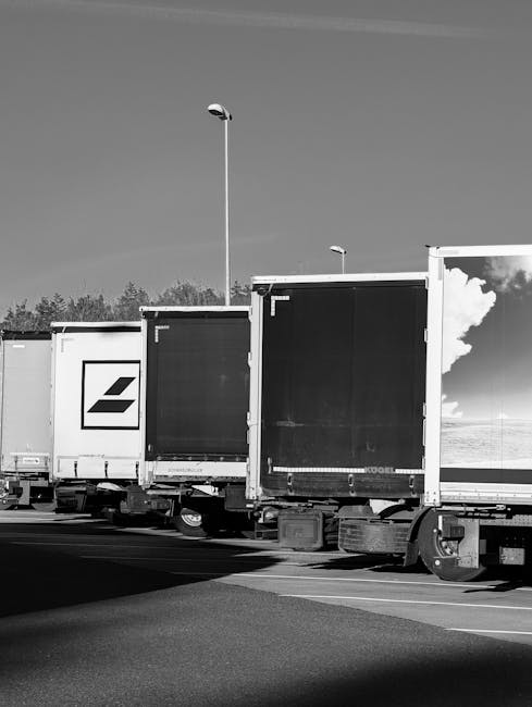 A row of large delivery trucks parked on a street with designated parking zones for house removals in Tottenham Hale. The trucks feature various company logos and are equipped with cargo doors, some of which are open or closed, indicating ongoing or prepared packing and loading processes. The rear area of the trucks is visible, showing mechanical wheels and undercarriage components, while the pavement and curb provide space for loading furniture, boxes, and packing materials such as cardboard boxes, protective blankets, and plastic wraps. Streetlights and trees are seen in the background under a clear sky, suggesting daytime conditions. This scene illustrates the logistics involved in home relocation, with trucks positioned for furniture transport and efficient loading, supporting the moving services offered by Man with Van Tottenham Hale, in compliance with parking restrictions and loading zones designated for removals teams.