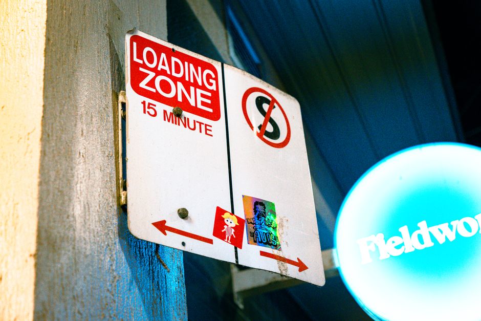 A rectangular metal sign mounted on a concrete wall indicating a loading zone with a red and white color scheme. The sign displays the words 'LOADING ZONE' and '15 MINUTE' in bold red letters, with a left-pointing arrow at the bottom left corner. To the right, there is a circular no-parking symbol with a red slash through a black 'P', and a character sticker positioned below the arrow. The sign appears slightly weathered with a few small stickers attached, including one colorful holographic sticker and a small red and yellow sticker. The surrounding environment suggests an urban setting, with part of a blue structure and a lit circular sign with the word 'Fieldwork' visible in the background. This scene likely relates to house removals and furniture transport, as the sign marks a designated area for loading and unloading during a home relocation, supported by services such as those provided by [COMPANY_NAME], Man with Van Tottenham Hale.