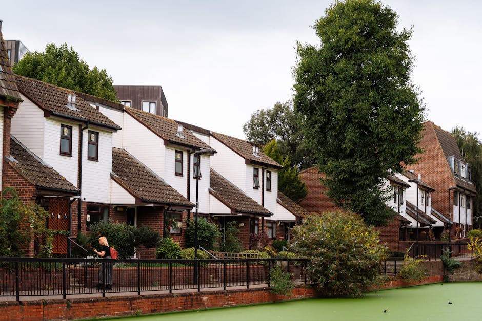 A residential street scene showing a row of modern houses with white cladding and brown tiled roofs, situated behind a black metal fence along a brick wall. In the foreground, a person with a black top and blonde hair is seen carrying a cardboard box, part of a home relocation or packing process. Behind them, there are additional cardboard boxes, plastic wrap, and packing materials stacked on the pavement indicating packing and moving activities. The background features trees with dense green foliage, and the sky appears overcast. The surroundings are well-maintained, with some shrubbery and small trees, and the area likely relates to house removals or furniture transport services provided by Man with Van Tottenham Hale, as seen in the context of the webpage about removals on Broadwater Farm Estate.