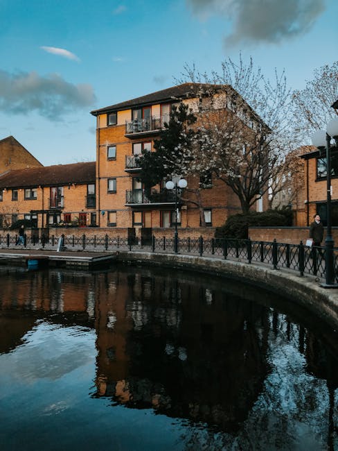 A multi-storey residential building constructed with orange brickwork, situated alongside a canal or waterway with calm, reflective water. The building has several small balconies with railings and patio furniture, and a pitched roof covered in dark shingles. In front of the property, there is a paved walkway bordered by a black metal fence, with a few people walking along it, some carrying small bags or parcels. A mature tree with sparse branches is present near the building, and a lamppost with multiple round lights is visible along the path. The sky above is partly cloudy with blue patches, and the scene appears to be taken during late afternoon or early evening. The setting suggests a peaceful residential area where property moving or furniture transport may be taking place, as part of house removals or home relocation services offered by Man with Van Tottenham Hale.