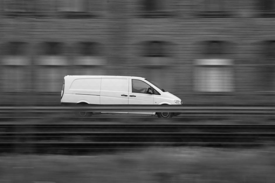 A white commercial van with a streamlined design is captured in motion on a motorway, with blurred horizontal lines indicating high speed. The van is moving across the frame from left to right, with a driver visible through the window. In the background, there is a brick wall with multiple small rectangular windows, which is also blurred due to the motion. The road surface and a metal barrier are visible below the van, contributing to the dynamic, fast-moving scene. This image reflects the logistical aspect of home relocation or furniture transport, as part of a professional removals service, highlighting efficient vehicle use in the loading process or moving logistics for residential or commercial moves in the N17 area.