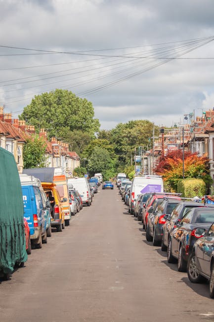 A quiet residential street during daytime with a straight asphalt road lined with parked cars on both sides, including sedans, hatchbacks, and vans, some of which are partially covered with protective tarps or covers. The street is bordered by terraced houses with brick facades, small front gardens, and a mix of deciduous trees with green and reddish foliage. Overhead, there are visible utility wires stretching across the sky, which is partly cloudy with patches of blue. In the distance, a man from Man with Van Tottenham Hale, a professional removals company, is seen loading or unloading furniture and cardboard boxes from a van parked at the curb, indicating a home relocation or moving process is underway. The overall scene captures the typical environment for furniture transport and packing during house removals in Tottenham Hale.