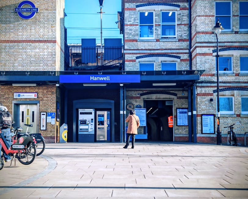 A woman dressed in a pink coat and dark trousers stands on the paved pavement outside Hanwell station, part of the Elizabeth Line, with her back turned towards the station entrance. The station features a brick façade with a dark blue awning displaying the name 'Hanwell' in white letters. To her left, there are two bike-sharing rental bikes, branded with Uber and VU, parked at bike stands. Next to the bikes, an automated ticket machine and a smaller ticket kiosk are visible, along with a yellow barrier indicating the lift access. The station entrance is flanked by two large digital information screens mounted on the brick wall, showing train schedules and service announcements. To the right of the entrance, a black bicycle is secured to a bike rack, and a tall streetlamp with a classic design is mounted on the corner of the building. The environment is well-lit with natural daylight, and some personnel in high-visibility jackets may be present, hinting at ongoing access or maintenance activities, relevant to local house removals and furniture transport services provided by Man with Van Tottenham Hale. The scene depicts a typical London train station's exterior, focusing on transport and logistics context for home relocation projects.