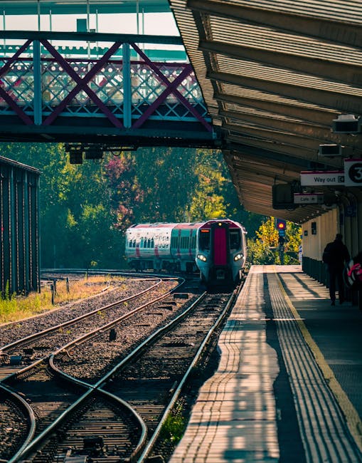 A modern train with a white and pink exterior approaching Tottenham Hale Station on railway tracks during daylight hours. The station platform on the right side of the image features a roof with corrugated metal panels and a tactile paving strip along the edge for safety. Several passengers are waiting near the platform, some standing close to the wall, while others are further along the platform. Overhead, a purple pedestrian bridge with metal railings and a lattice design connects the station platforms, allowing pedestrians to cross over the tracks. The background reveals trees with green foliage, indicating a pleasant day. This scene captures elements typical of home relocation logistics, such as transport via train, and is associated with professional removal services like Man with Van Tottenham Hale. The image emphasizes the process of furniture transport and the structural environment of a busy suburban station, relevant to moving and packing activities.'