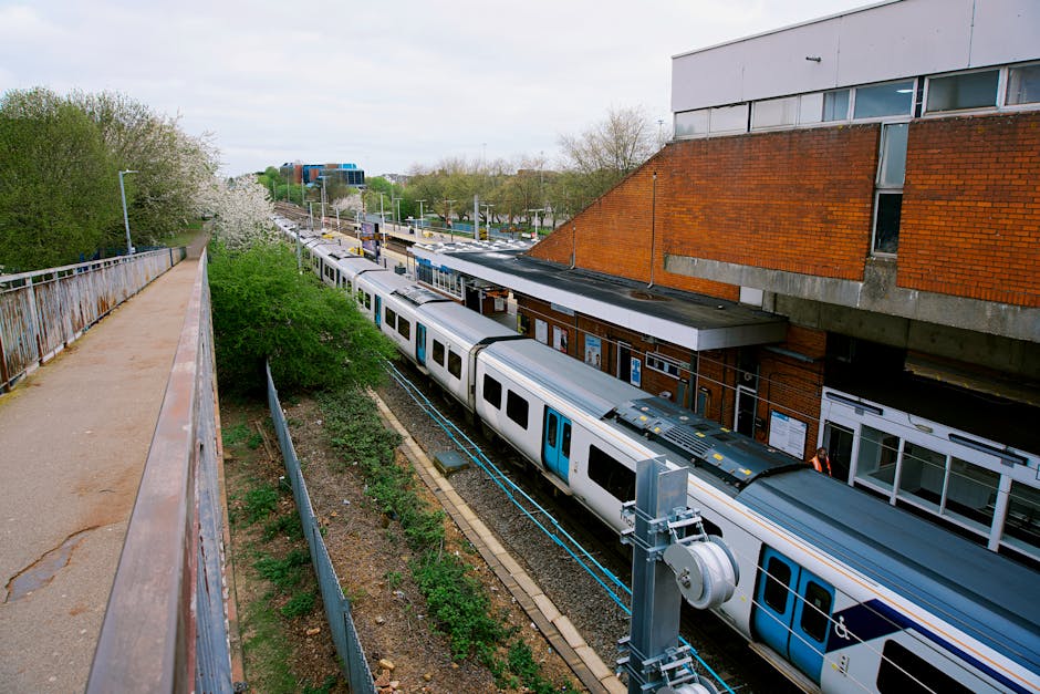 A woman dressed in a pink coat and dark trousers stands on the paved pavement outside Hanwell station, part of the Elizabeth Line, with her back turned towards the station entrance. The station features a brick façade with a dark blue awning displaying the name 'Hanwell' in white letters. To her left, there are two bike-sharing rental bikes, branded with Uber and VU, parked at bike stands. Next to the bikes, an automated ticket machine and a smaller ticket kiosk are visible, along with a yellow barrier indicating the lift access. The station entrance is flanked by two large digital information screens mounted on the brick wall, showing train schedules and service announcements. To the right of the entrance, a black bicycle is secured to a bike rack, and a tall streetlamp with a classic design is mounted on the corner of the building. The environment is well-lit with natural daylight, and some personnel in high-visibility jackets may be present, hinting at ongoing access or maintenance activities, relevant to local house removals and furniture transport services provided by Man with Van Tottenham Hale. The scene depicts a typical London train station's exterior, focusing on transport and logistics context for home relocation projects.